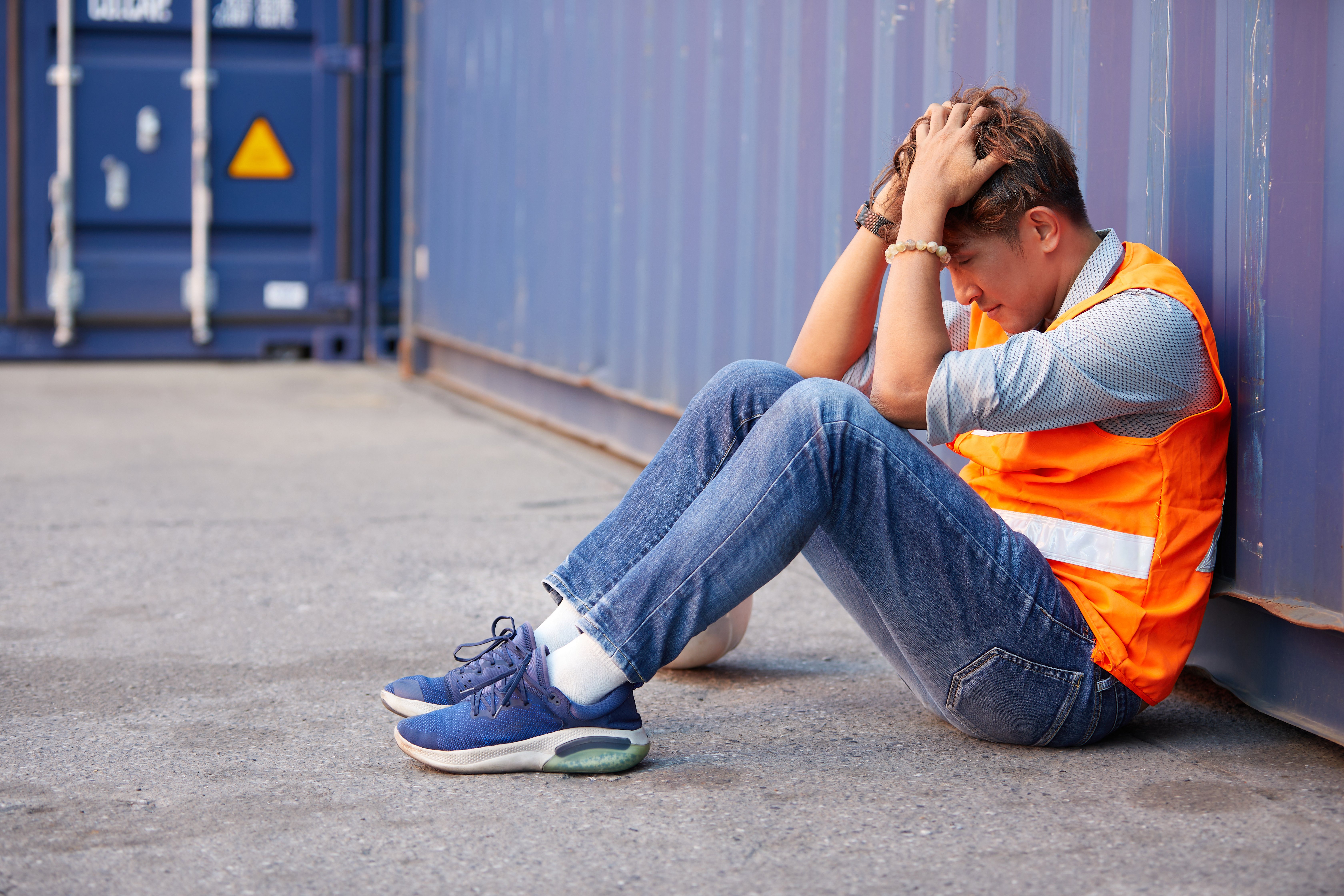 unhappy warehouse worker in orange vest sitting on ground leaning against shipping container after overtime at factory